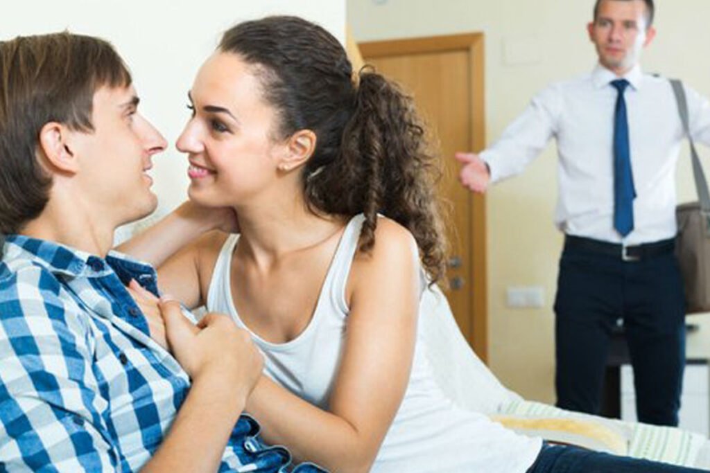 A man stands behind a woman gazing her as she is sitting on lap of another man.