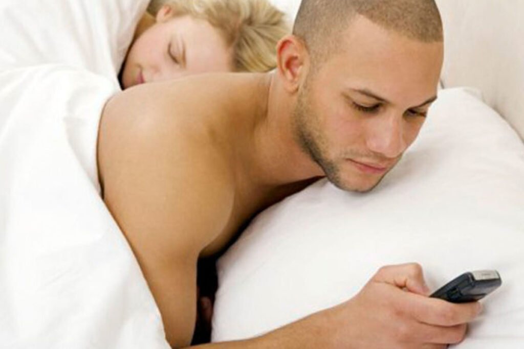 A couple lying in bed, husband engrossed in his cell phone.