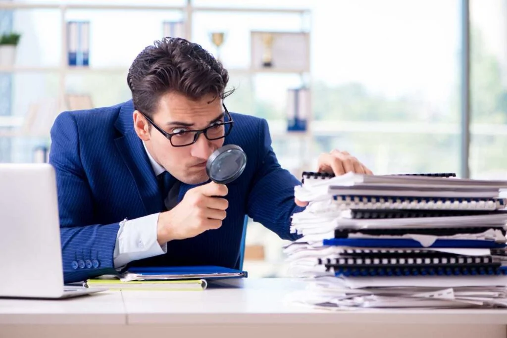 A man in a suit examines a stack of papers using a magnifying glass, searching for details.